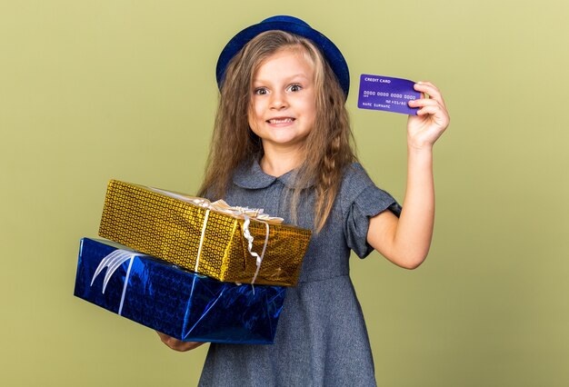 コピースペースでオリーブグリーンの壁に分離されたギフトボックスとクレジットカードを保持している青いパーティーハットで驚いた小さなブロンドの女の子