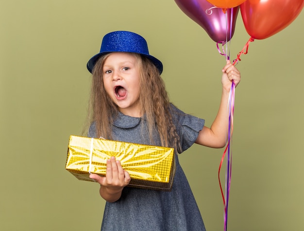 Bambina bionda sorpresa con cappello da festa blu che tiene palloncini di elio e confezione regalo isolato sulla parete verde oliva con spazio di copia