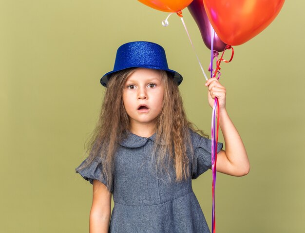 surprised little blonde girl with blue party hat holding helium balloons isolated on olive green wall with copy space