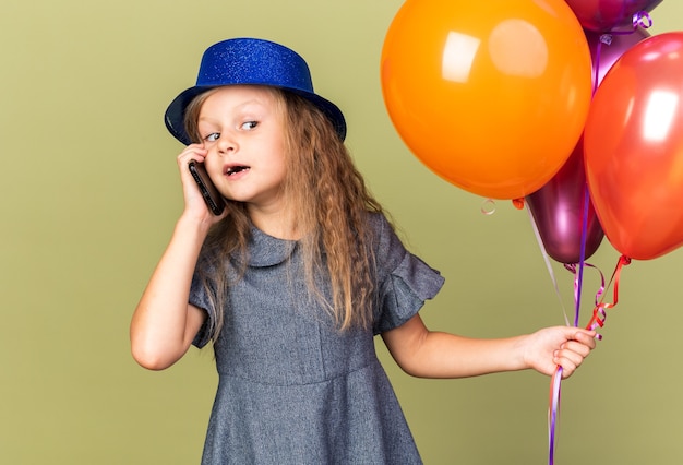 Sorpresa bimba bionda con cappello da festa blu che tiene palloncini di elio e parla al telefono guardando il lato isolato sulla parete verde oliva con spazio di copia