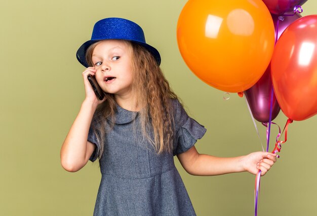 surprised little blonde girl with blue party hat holding helium balloons and talking on phone looking at side isolated on olive green wall with copy space