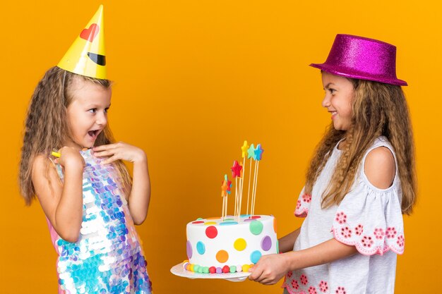 コピースペースでオレンジ色の壁に分離されたバースデーケーキを保持している紫色のパーティーハットを持つ小さな白人の女の子を見てパーティーキャップを持つ驚いた小さなブロンドの女の子