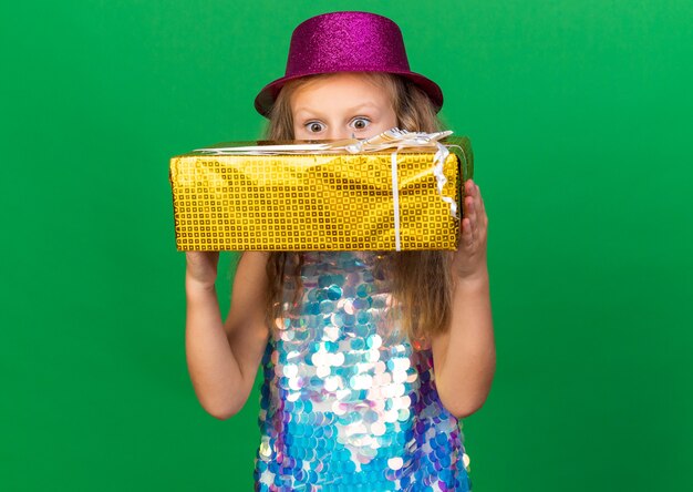 복사 공간 녹색 벽에 고립 된 선물 상자를 들고 보라색 파티 모자와 놀란 된 작은 금발 소녀