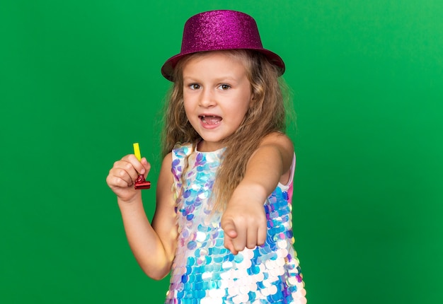 surprised little blonde girl with purple party hat holding party whistle and pointing  isolated on green wall with copy space