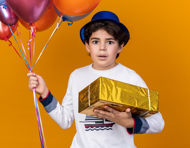 Surprised little boy wearing blue party hat holding balloons with gift box isolated on orange wall