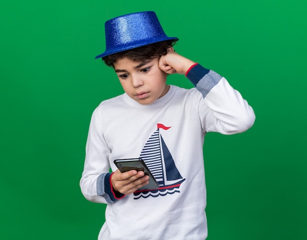 Удивленный маленький мальчик в синей партийной шляпе, держащий и смотрящий на телефон, изолированный на зеленой стене