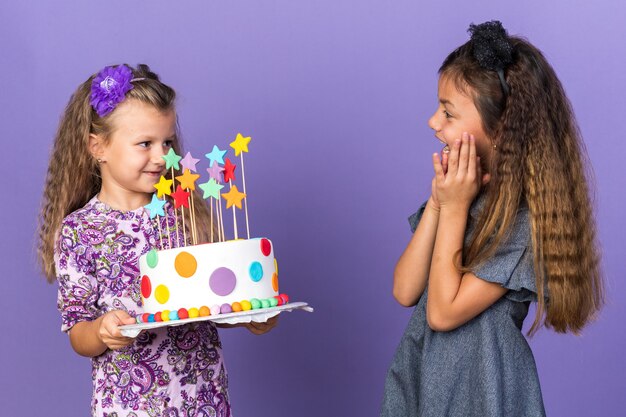 surprised little caucasian girl looking at pleased little blonde girl holding birthday cake isolated on purple wall with copy space
