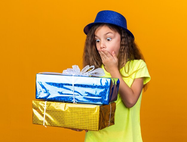 surprised little caucasian girl with blue party hat putting hand on mouth and holding gift boxes isolated on orange wall with copy space