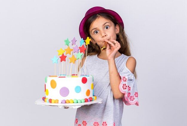 удивленная маленькая кавказская девушка в фиолетовой шляпе с праздничным тортом и свистком на белой стене с копией пространства