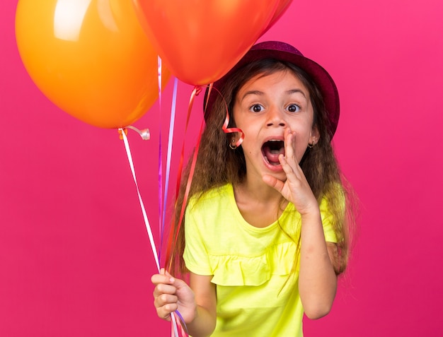 Piccola ragazza caucasica sorpresa con cappello da festa viola che tiene palloncini di elio e tiene la mano vicino alla bocca chiamando qualcuno isolato sul muro rosa con spazio di copia