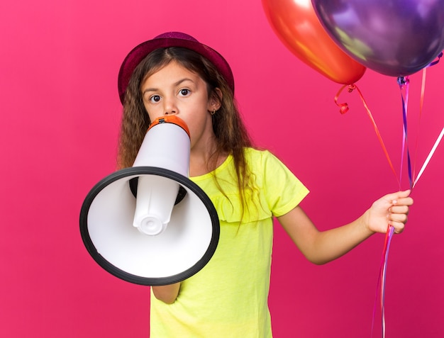 surprised little caucasian girl with purple party hat holding helium balloons and loud speaker isolated on pink wall with copy space