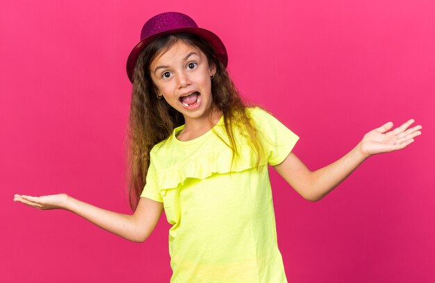 surprised little caucasian girl with purple party hat keeping hands open isolated on pink wall with copy space