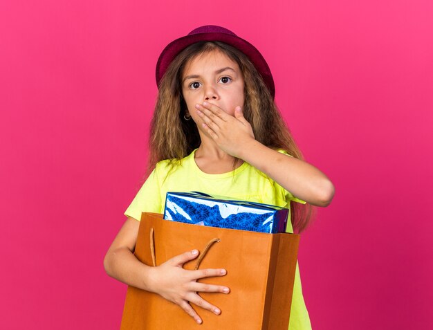 surprised little caucasian girl with purple party hat putting hand on mouth and holding gift box in paper bag isolated on pink wall with copy space