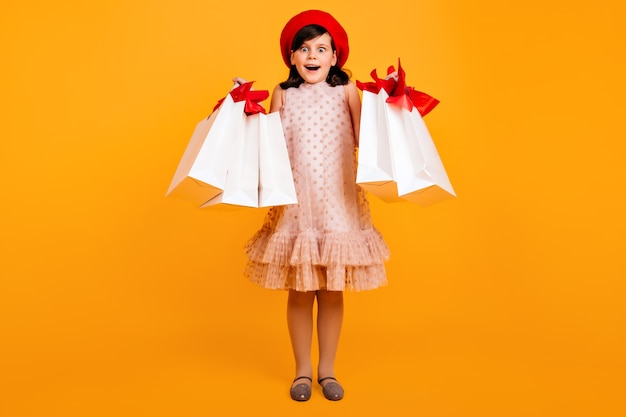 무료 사진 프랑스 베레모 쇼핑 후 포즈에 놀란 된 어린 소녀. 종이 봉투와 함께 놀란 아이.