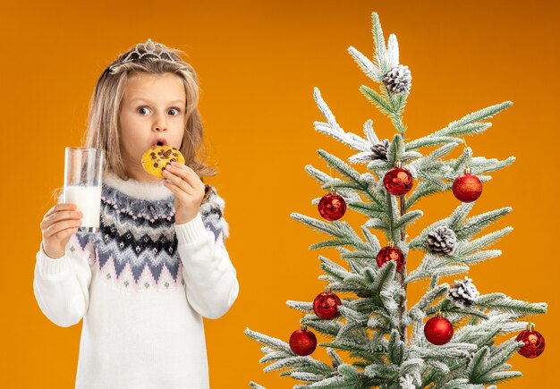 Surprised little girl standing nearby christmas tree wearing tiara with garland on neck holding glass of milk trying cookies isolated on orange background