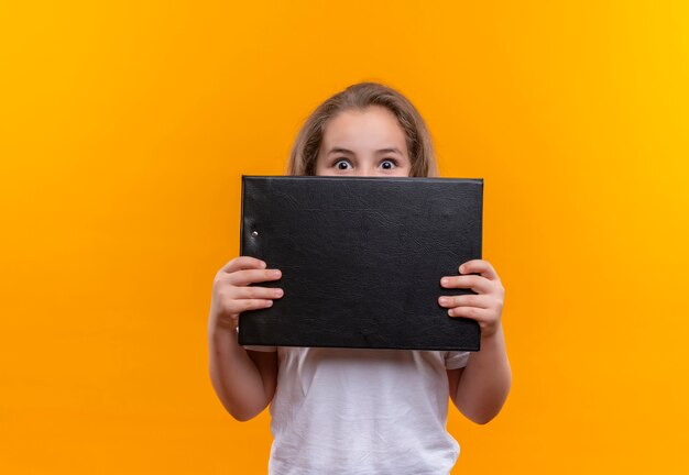 Surprised little school girl wearing white t-shirt covered mouth with clipboard on isolated orange background