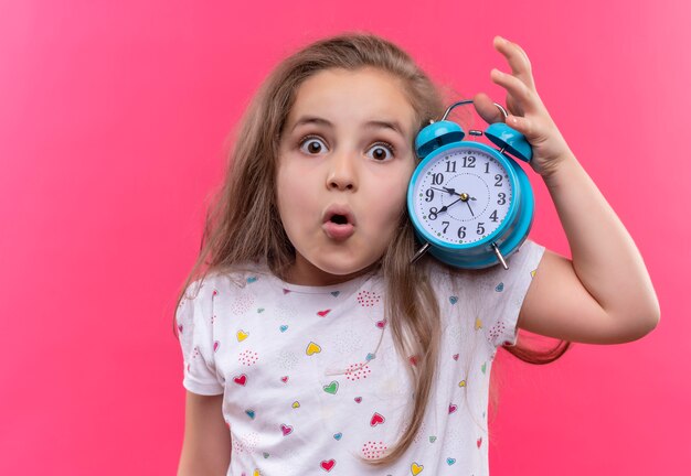 Surprised little school girl wearing white t-shirt holding alarm clock on isolated pink background