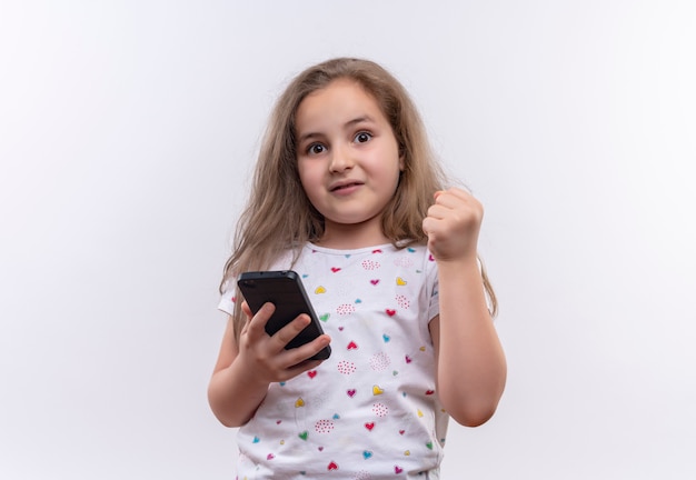 Piccola ragazza sorpresa della scuola che indossa il telefono bianco della tenuta della maglietta e pugno di sollevamento su fondo bianco isolato