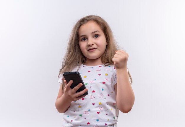 Удивленная маленькая школьница в белой футболке держит телефон и поднимает кулак на изолированном белом фоне