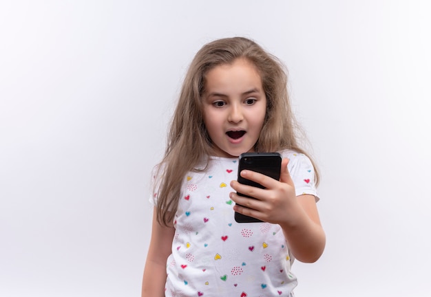 Surprised little school girl wearing white t-shirt looking on phone on isolated white background