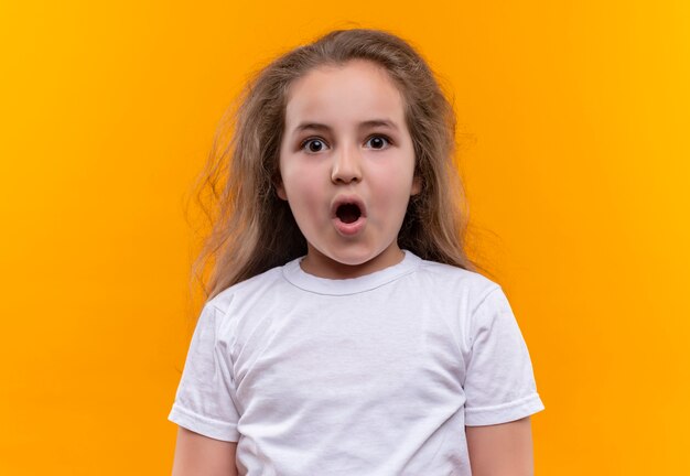 Удивленная маленькая школьница в белой футболке, открывающая рот на изолированном оранжевом фоне