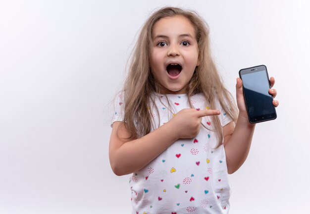 Surprised little school girl wearing white t-shirt points to phone on isolated white background