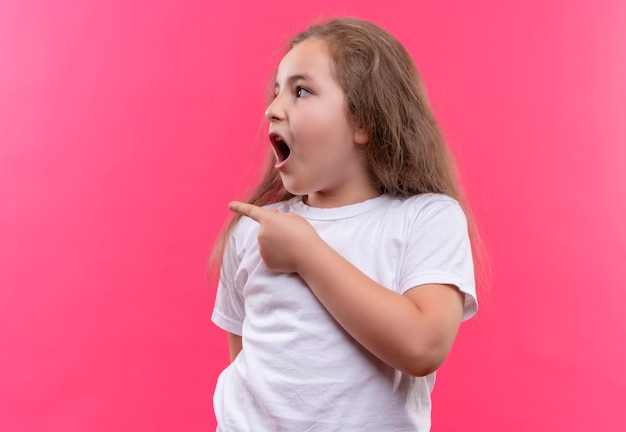 白いTシャツを着て驚いた小さな女子高生は、孤立したピンクの背景に横を指しています