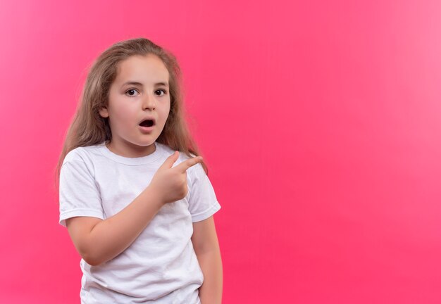Surprised little school girl wearing white t-shirt points to side on isolated pink background