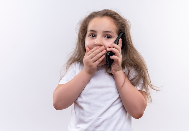 Surprised little school girl wearing white t-shirt speaks on phone covered mouth on isolated white background