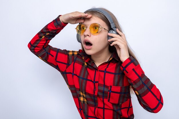 Surprised looking at distance with hand beautiful little girl wearing red shirt and glasses with headphones 