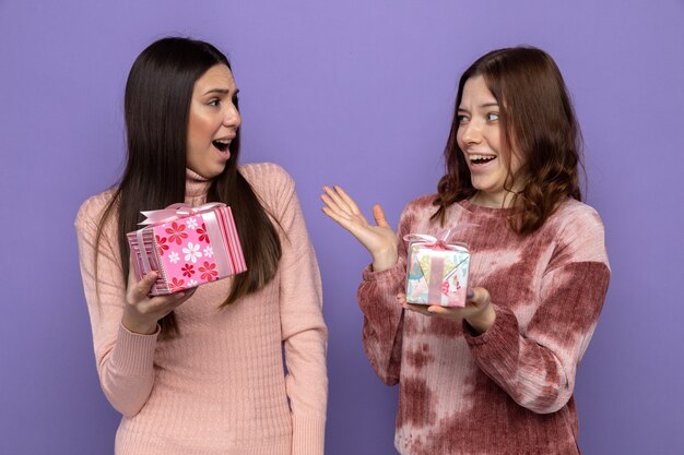 Surprised looking at each other two girls on happy woman's day holding presents isolated on blue wall