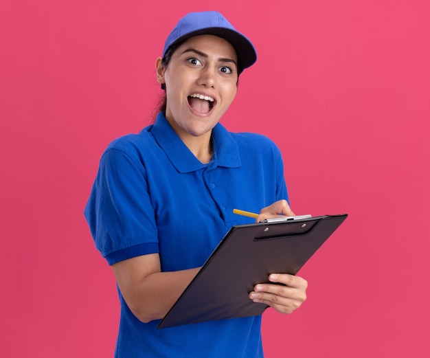 Surprised looking at front young delivery girl wearing uniform with cap writing something on clipboard isolated on pink wall