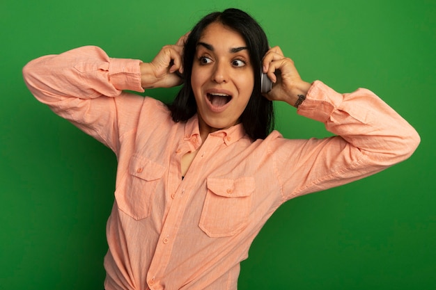 Foto gratuita sorpreso guardando lato giovane bella ragazza che indossa la maglietta rosa con le cuffie isolate sulla parete verde