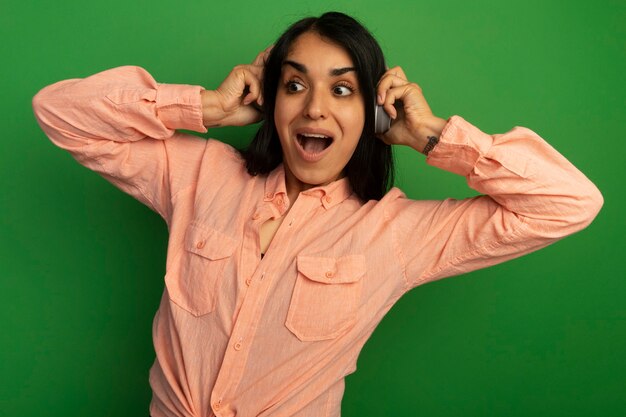 Surprised looking at side young beautiful girl wearing pink t-shirt with headphones isolated on green wall