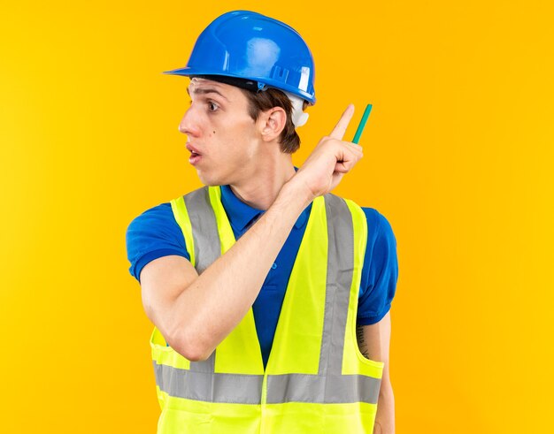 Surprised looking side young builder man in uniform holding pencil and points at behind isolated on yellow wall with copy space