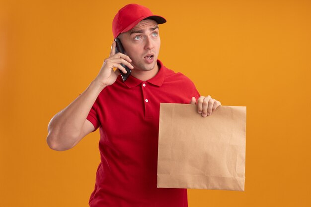 Удивленный, глядя в сторону, молодой курьер в униформе с кепкой, держащей бумажный пакет с едой, разговаривает по телефону, изолированному на оранжевой стене