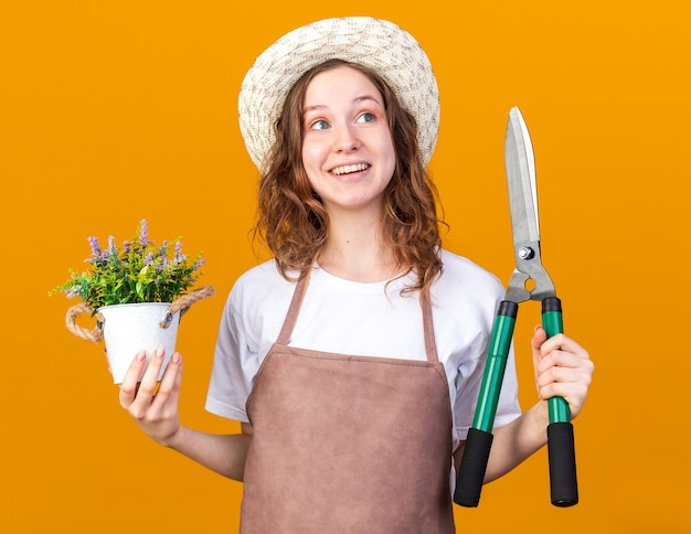 Surprised looking side young female gardener wearing gardening hat holding flower in flowerpot with pruning shear 