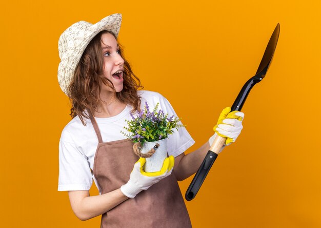 Surprised looking side young female gardener wearing gardening hat holding flower in flowerpot with spade 