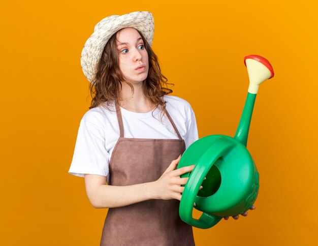 Surprised looking side young female gardener wearing gardening hat holding watering can isolated on orange wall
