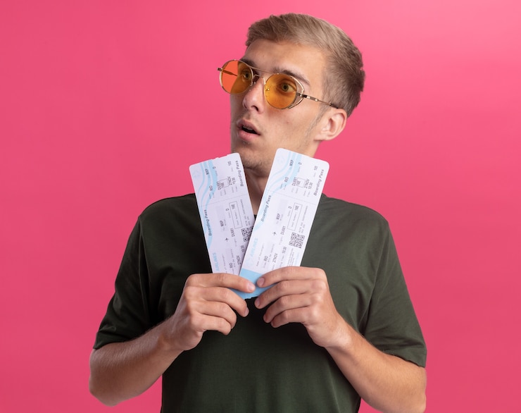 Free Photo surprised looking at side young handsome guy wearing green shirt and glasses holding tickets isolated on pink wall