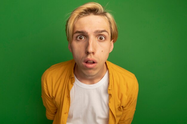 Surprised looking straight ahead young blonde guy wearing yellow t-shirt isolated on green
