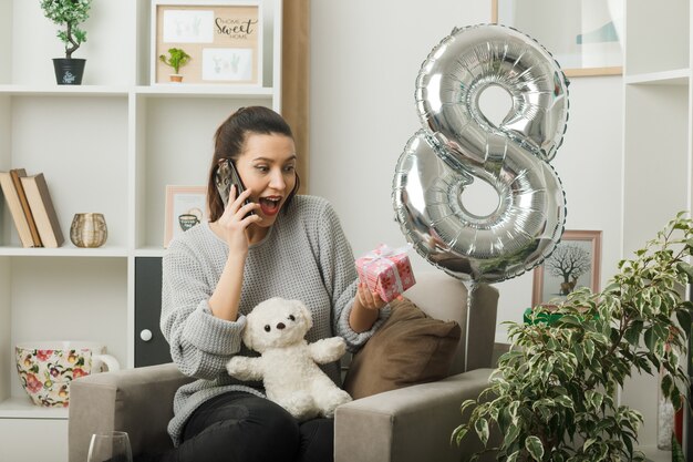 Surprised looking up beautiful girl on happy women day holding present speaks on phone sitting on armchair in living room