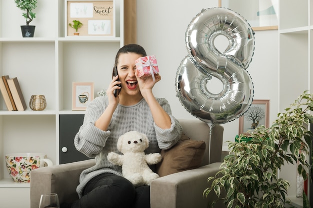 Surprised looking up beautiful girl on happy women day holding present speaks on phone sitting on armchair in living room