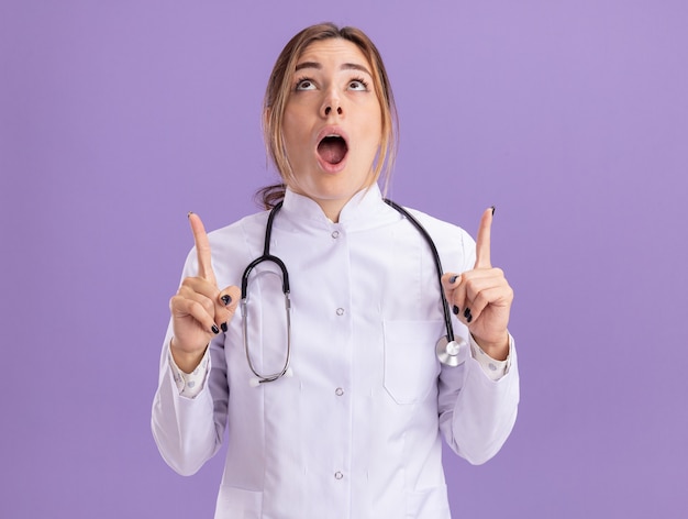 Surprised looking up young female doctor wearing medical robe with stethoscope points at up isolated on purple wall