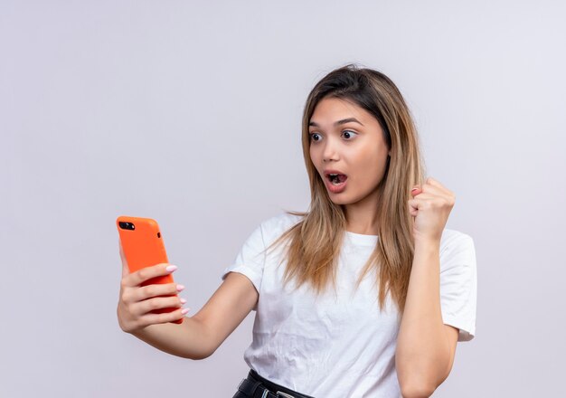 Удивленная милая молодая женщина в белой футболке смотрит на мобильный телефон, поднимая сжатый кулак на белой стене