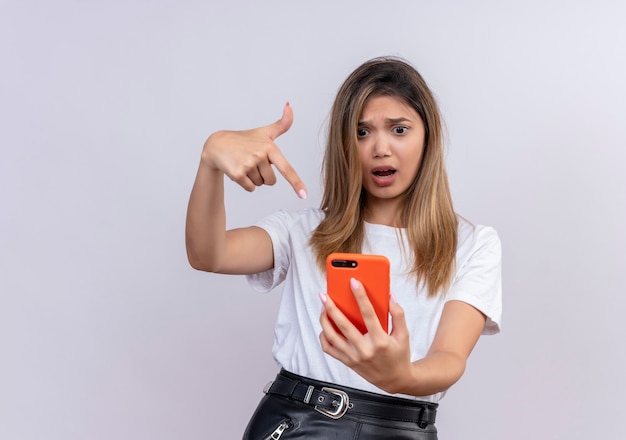 A surprised lovely young woman in white t-shirt pointing with index finger while looking at mobile phone on a white wall