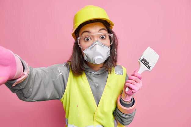 Удивленная работница по техобслуживанию носит униформу защитной маски и в очках фотографирует себя, держит кисть, использует строительные инструменты для ремонта.