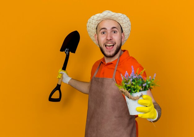 Surprised male gardener wearing gardening hat and gloves holding spade and flowers in flowerpot 