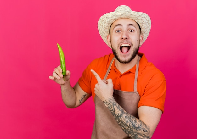 Surprised male gardener wearing gardening hat holds and points at hot pepper