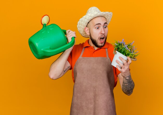 Surprised male gardener wearing gardening hat holds watering can and flowers